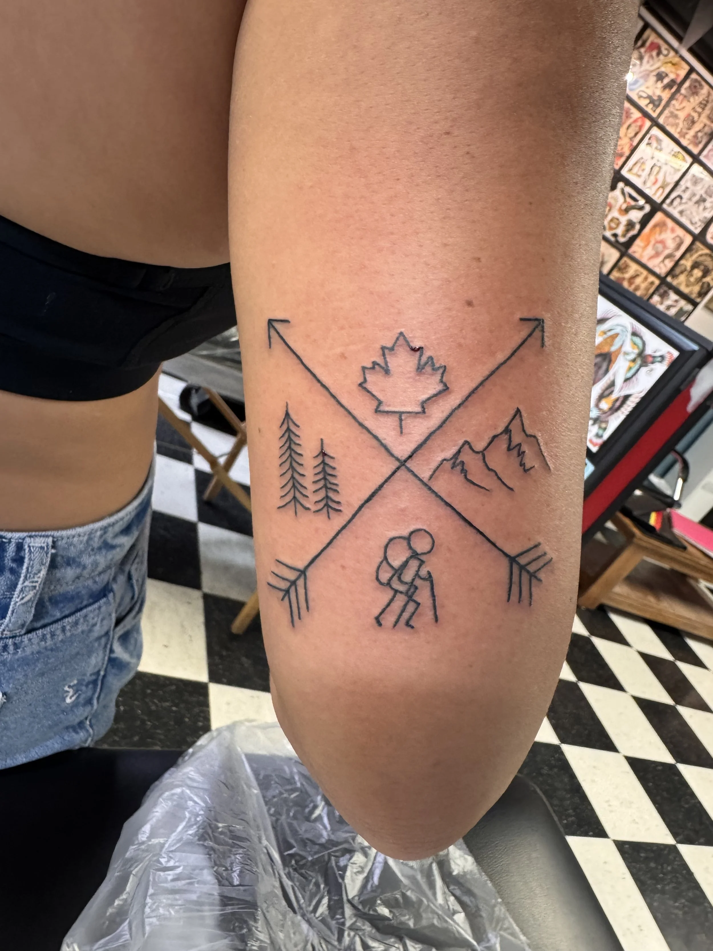 A close-up view of a person's arm with a tattoo of a maple leaf and mountain peaks, with black outline and intersecting arrows, set in a tattoo shop with tattoo flash artwork on the wall.