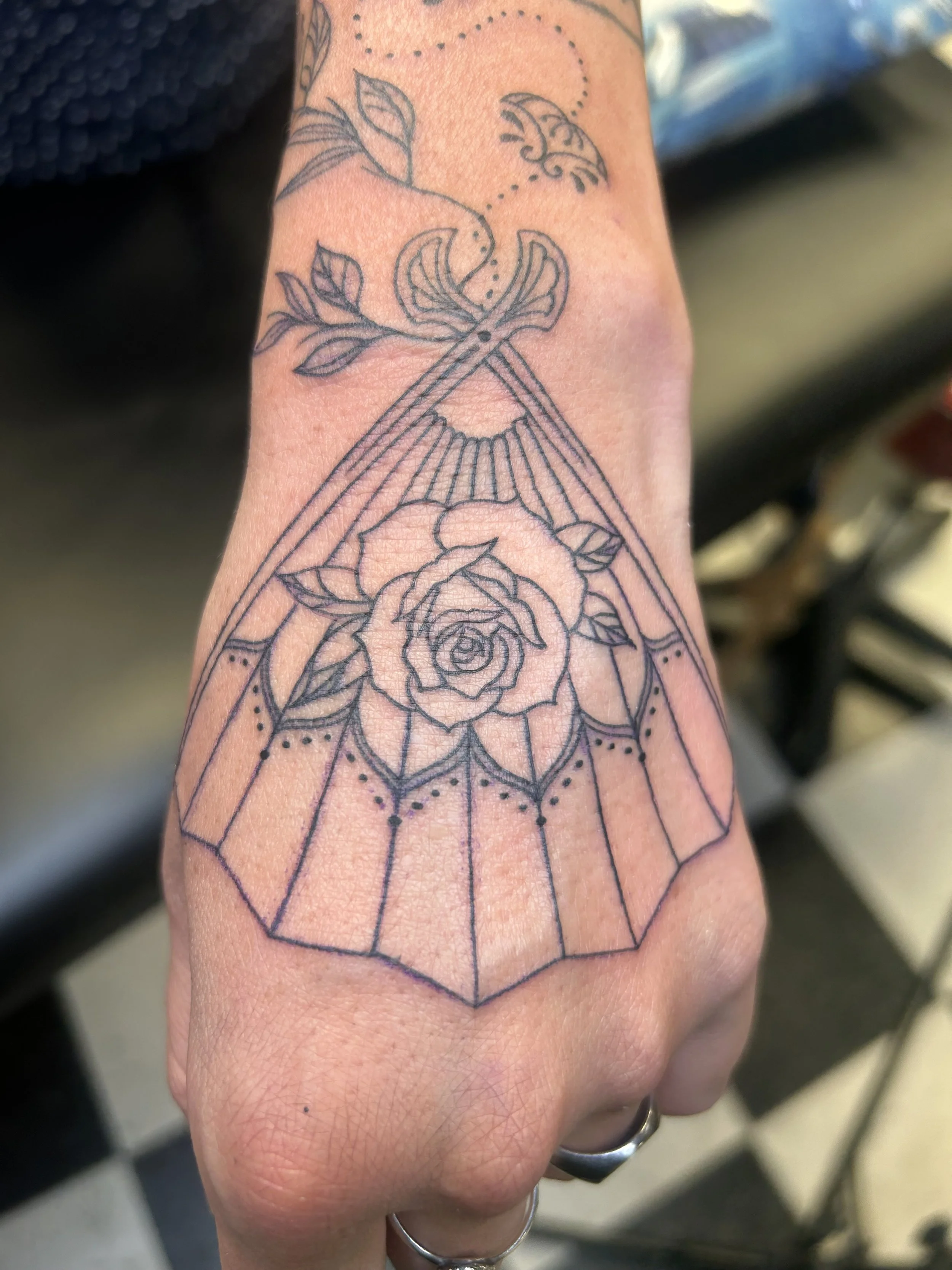 A tattoo of a rose within a geometric shape with leaves and dotted line details on a person's wrist.