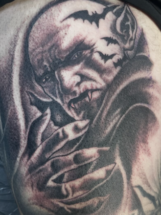 Close-up of a detailed black and gray tattoo of a ghoulish, scary face with sharp teeth, a forest bat, and long fingernails.