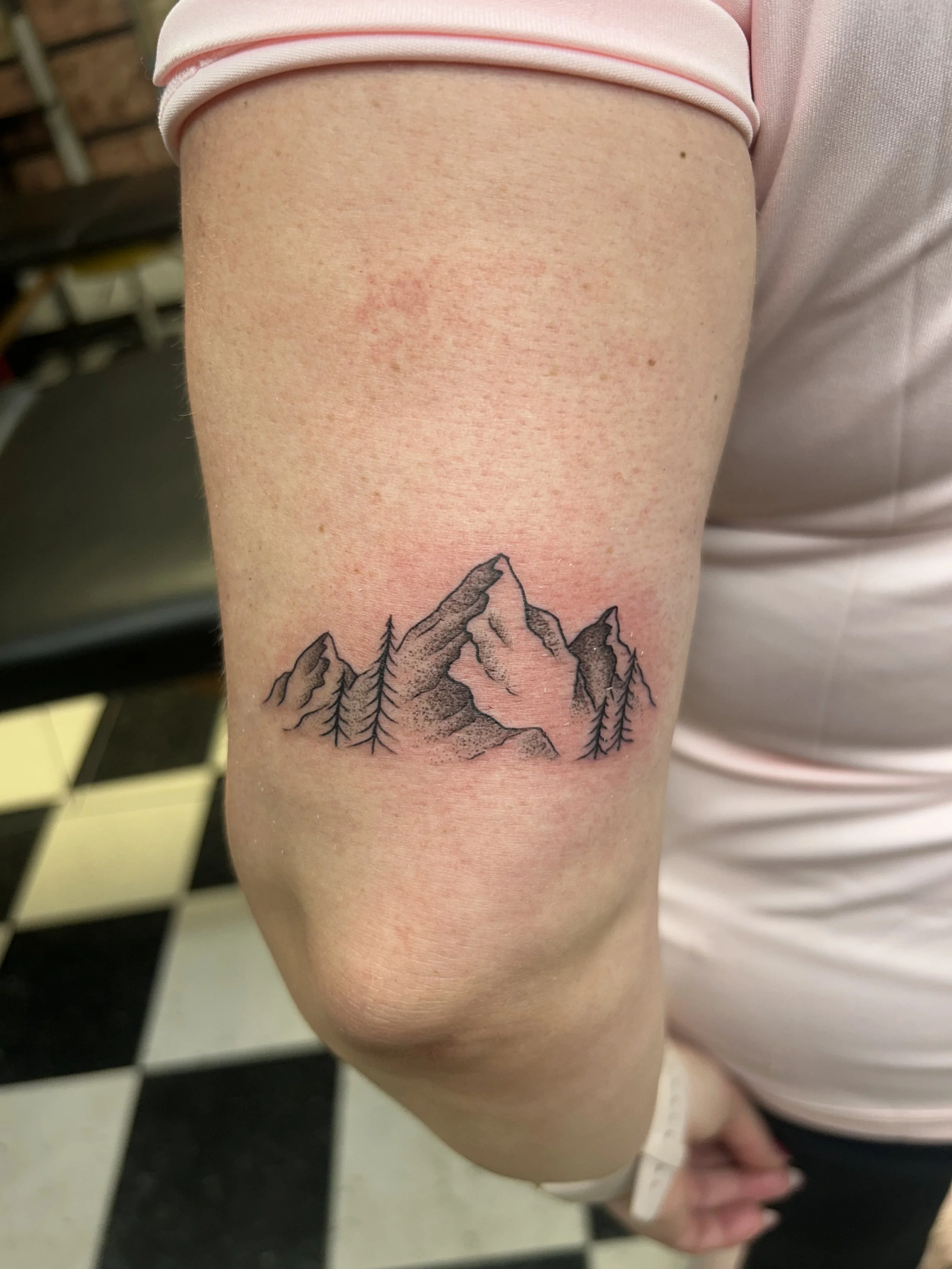 Tattoo of mountains and pine trees on a person's upper arm.