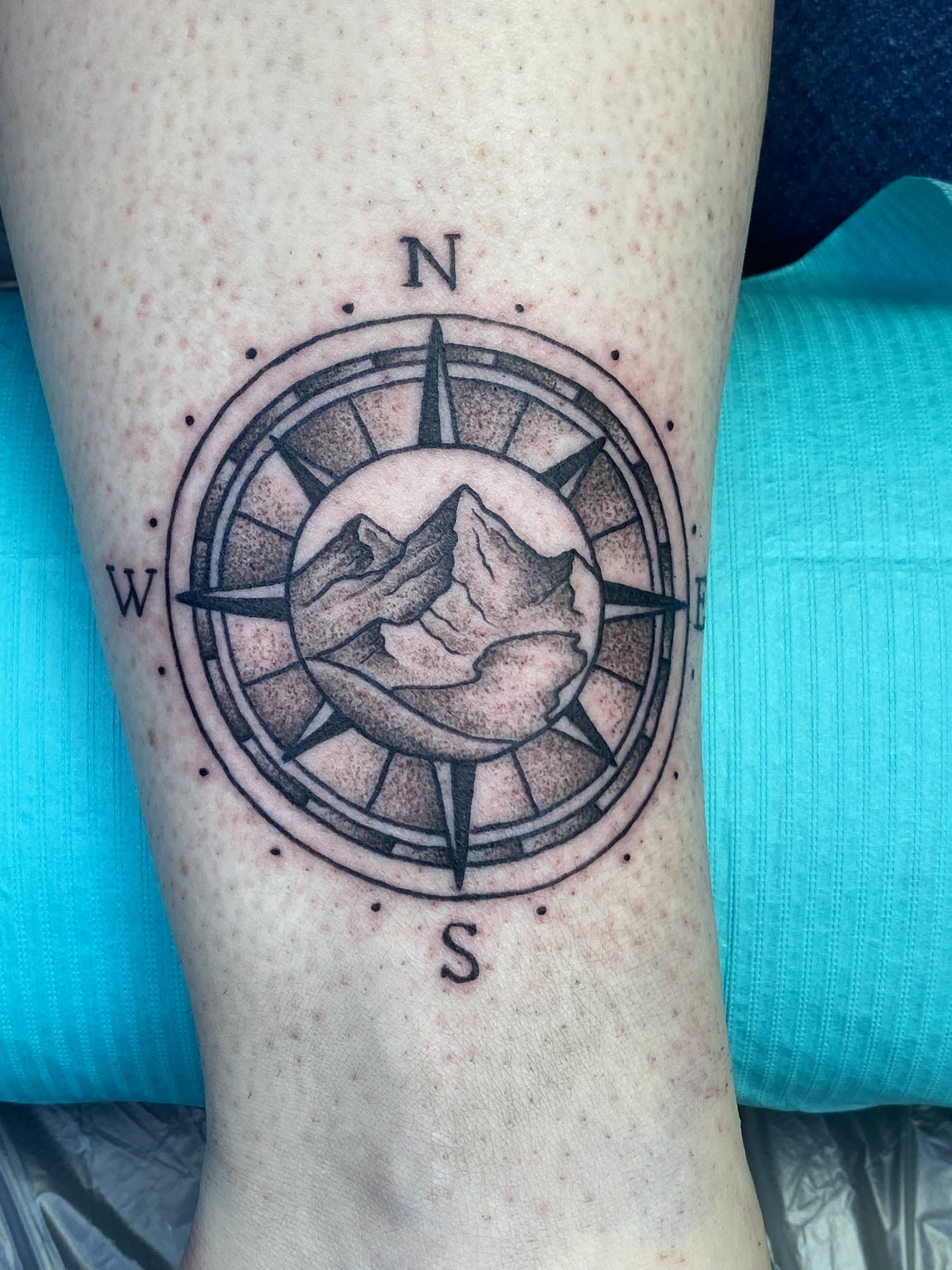 A tattoo of a compass with mountain scenery inside the circular part, with the cardinal directions N, S, W, E marked around it, on a person's thigh.