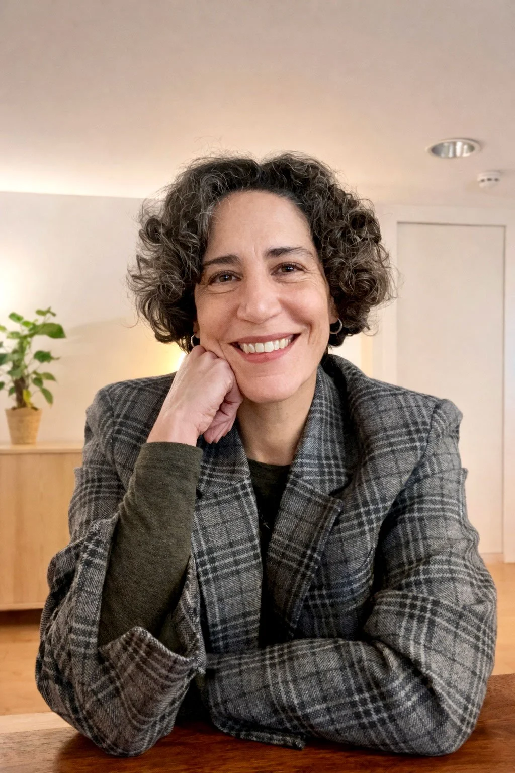 A woman with curly dark hair smiling at the camera, resting her face on her hand, wearing a plaid blazer, sitting at a wooden table in a cozy, well-lit room with a plant on a cabinet in the background.