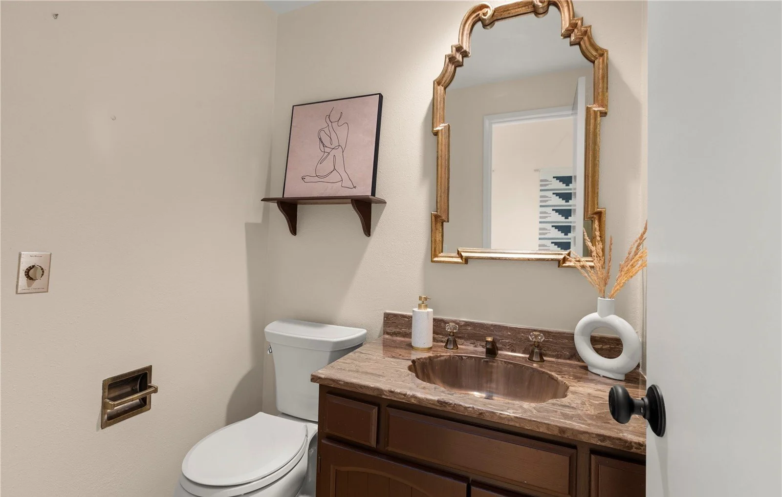 A small bathroom with a white toilet, a wooden vanity with a brown marble countertop and sink, a gold framed mirror, a white vase with dried plants, a soap dispenser, a wall shelf with artwork, and a partial view of a door.