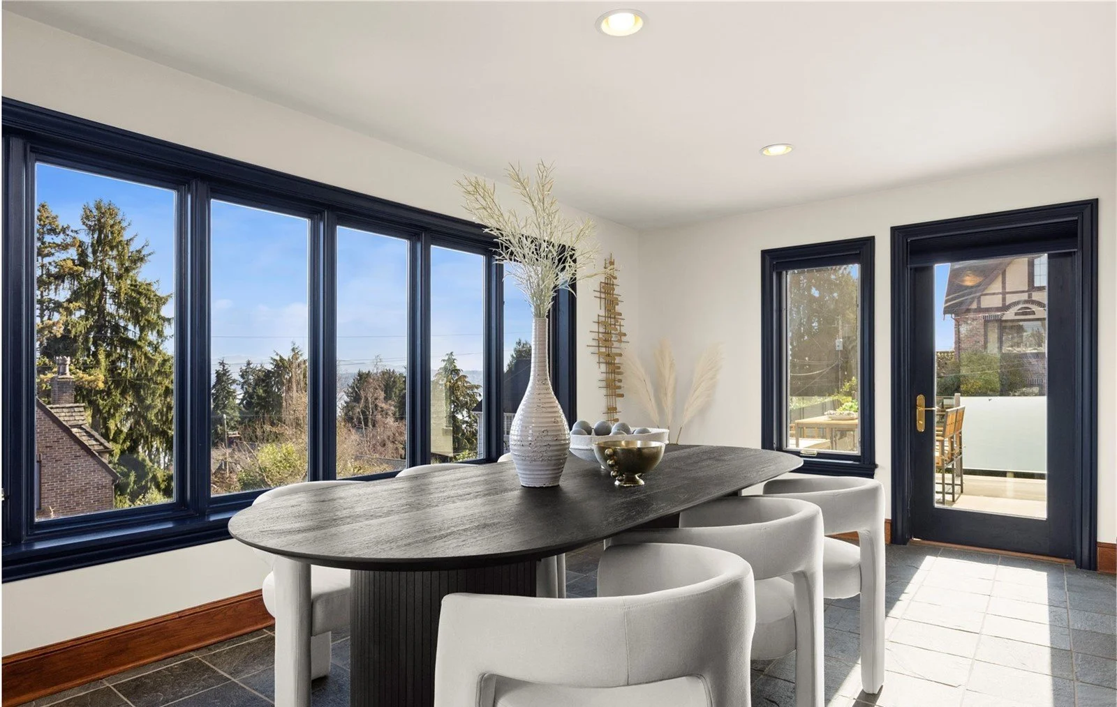 Bright dining room with large windows showing outdoor trees and blue sky, white walls, dark wood door with glass panel, and a dark wood dining table with five white chairs, decorated with vases and bowls, and a balcony with outdoor seating.