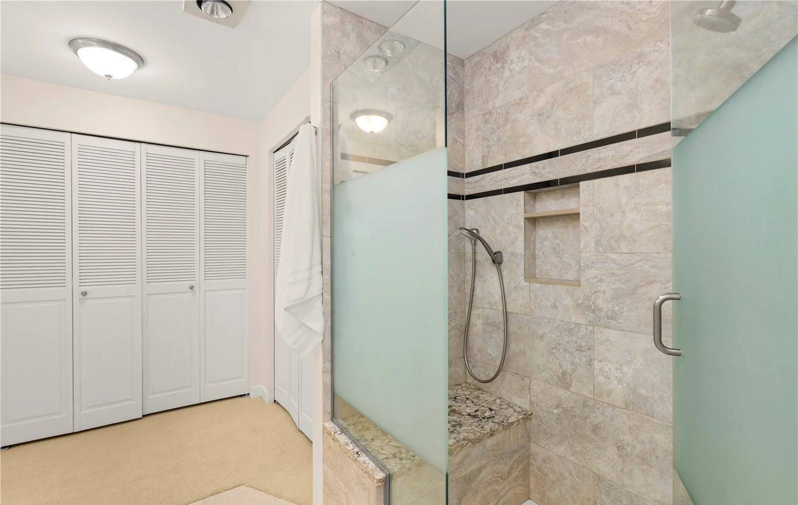 Bathroom with a linen closet, a white towel hanging on a hook, and a walk-in shower with beige tiles and a black decorative stripe.