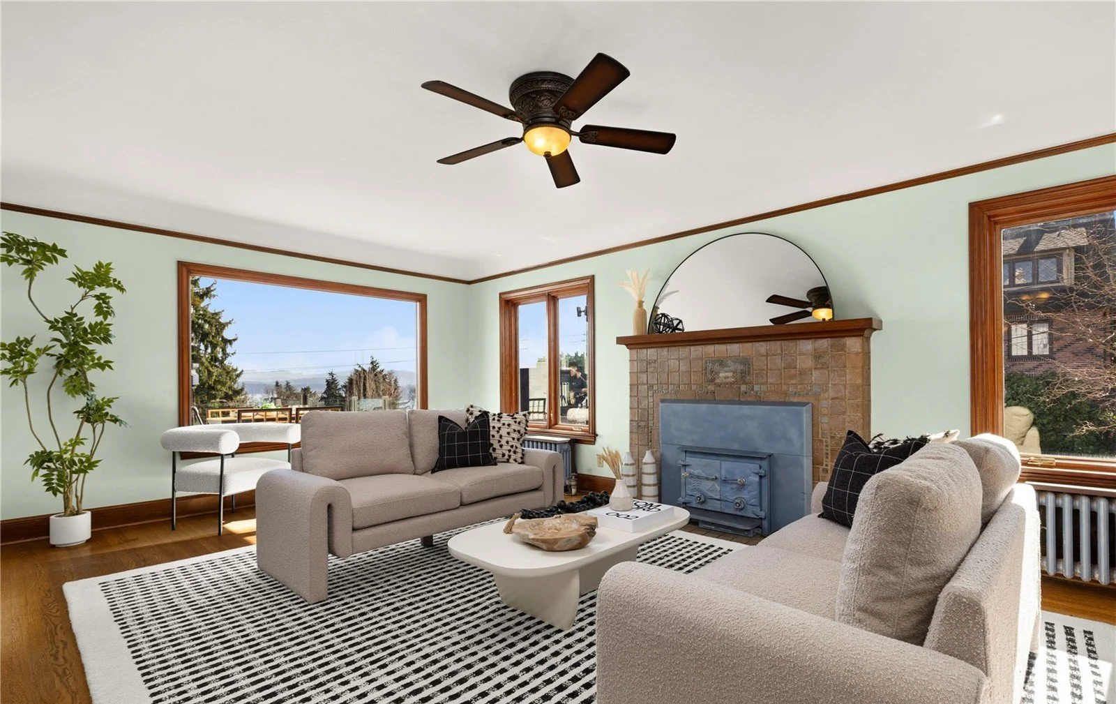 Living room with two beige sofas, a white coffee table, a black-and-white patterned rug, large windows showing outdoor views, a brick fireplace with a round mirror above, and a ceiling fan.