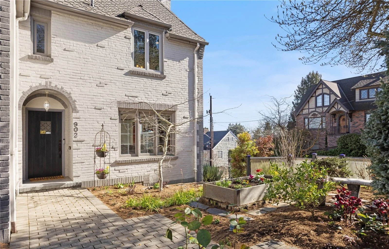 Front view of a white brick house with black front door, small windows, and a garden with plants and trees.