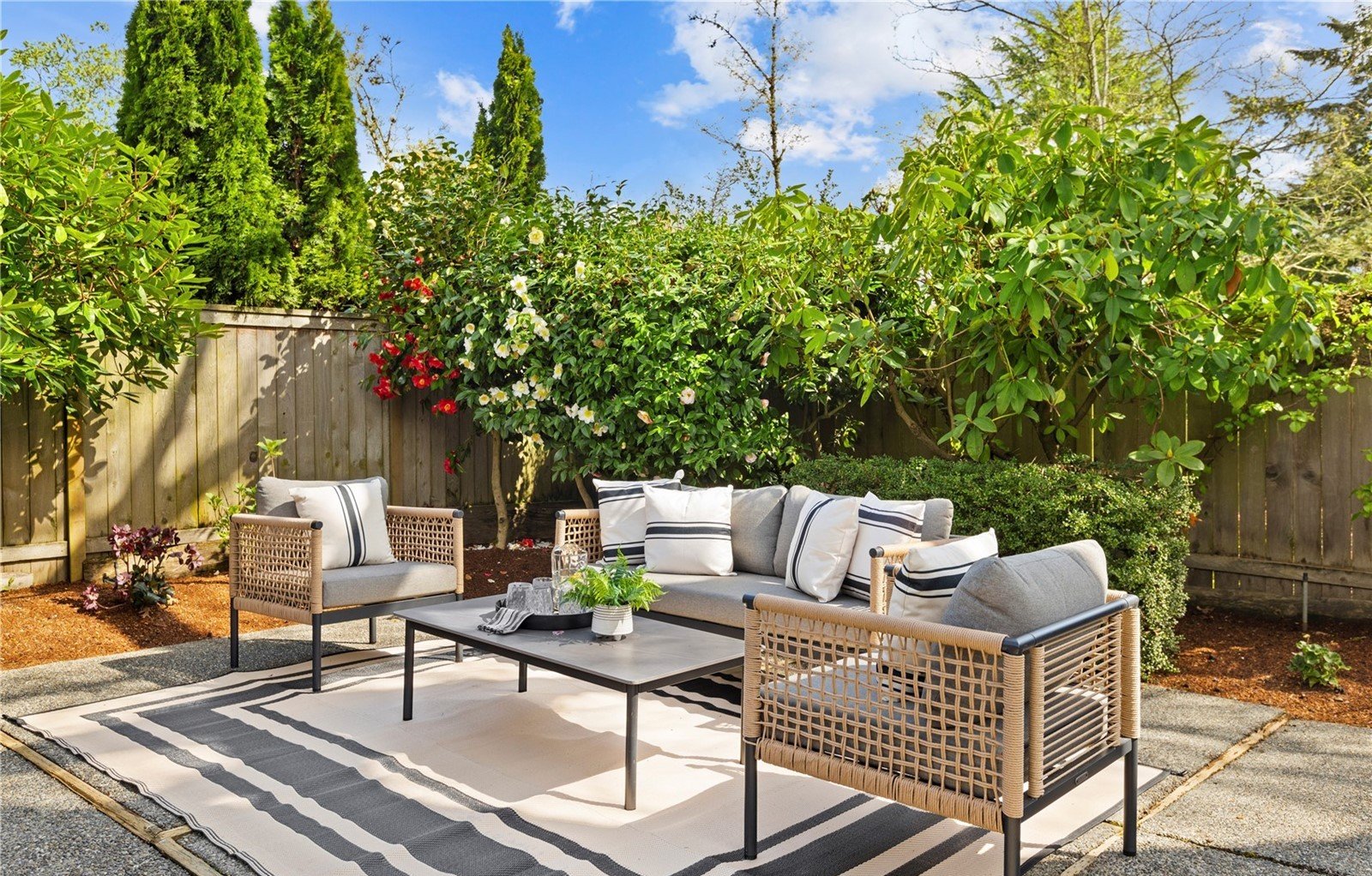 Outdoor patio with woven rattan style sofas and a coffee table on a black and white striped rug, surrounded by green bushes and trees with a wooden fence in the background, under a blue sky with some clouds.