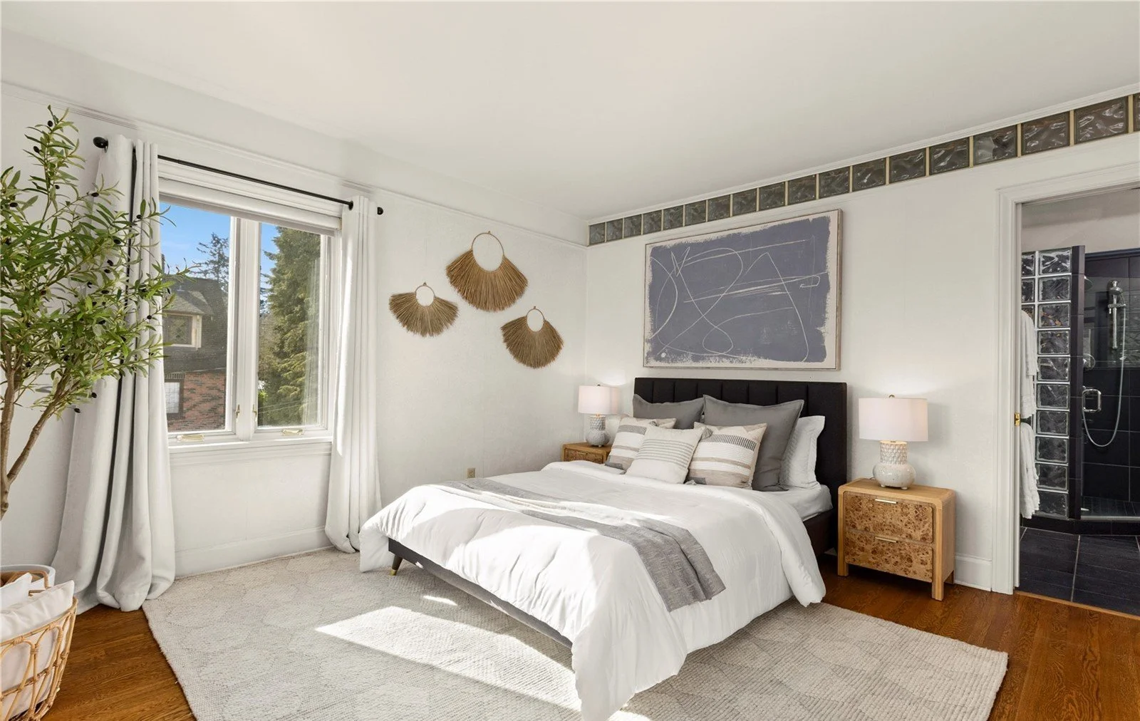 Bright bedroom with white walls, large window with white curtains, bed with gray and white pillows, two wooden nightstands with lamps, abstract art on the wall, and a doorway leading to a black-tiled bathroom with a shower.