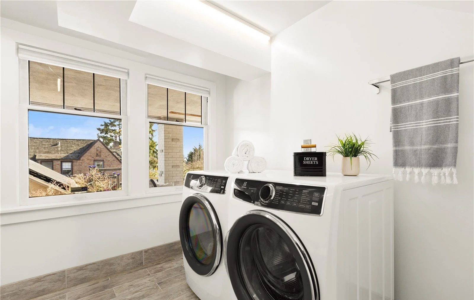 A laundry room with a front-loading washer and dryer next to a window, with neatly folded towels on top and a potted plant, a black box labeled "Dryer Sheets," a small decorative box, and a gray towel hanging on a bar on the wall.