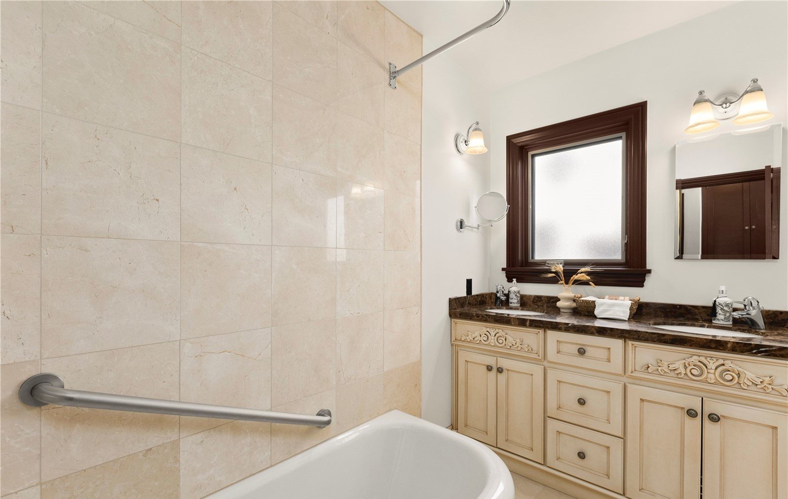 Bathroom with a bathtub, beige tiled wall, dark wood framed window, cream-colored vanity with dark brown marble countertop, double sink, and two wall-mounted light fixtures.