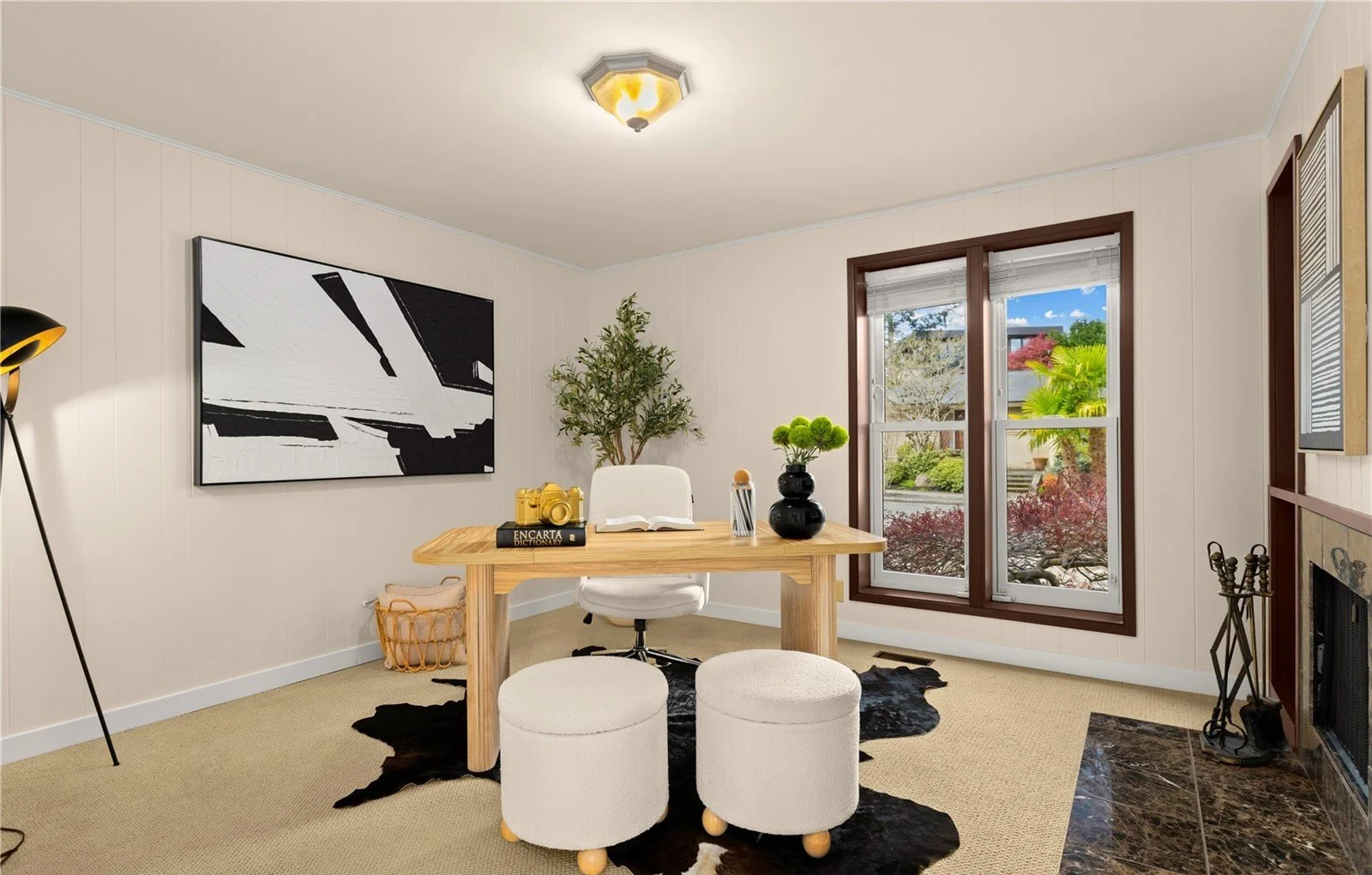 Home office with wooden desk, white swivel chair, cowhide rug, large abstract black and white painting, window with a garden view, small potted plants, and fireplace.