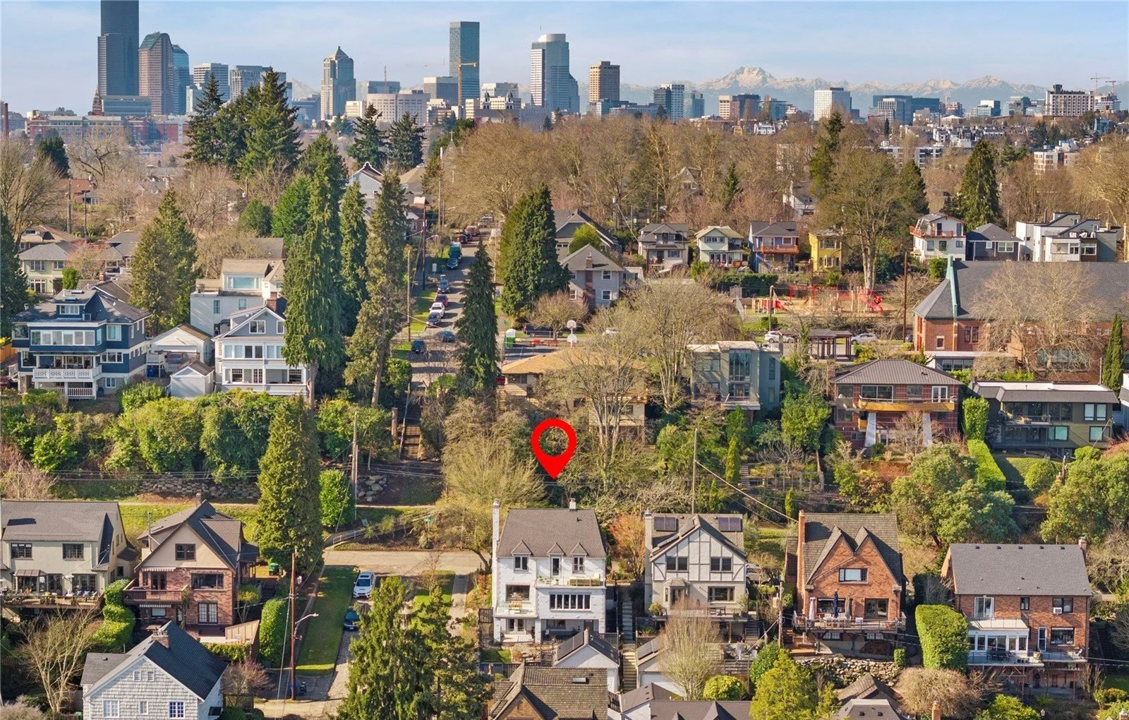Aerial view of a residential neighborhood with various houses, trees, and streets, with a city skyline in the background and mountains in the distance.