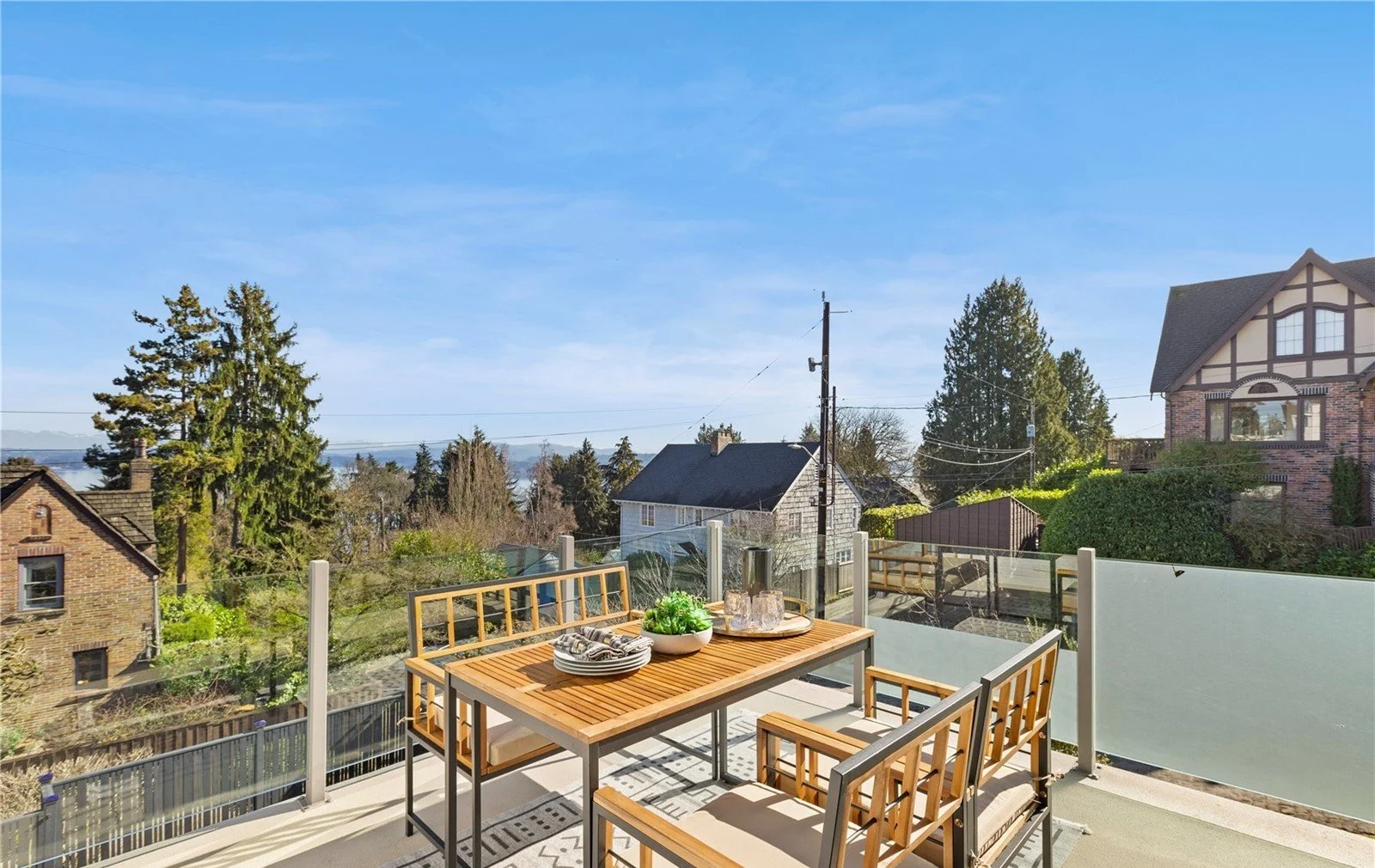 Outdoor balcony with wooden table and chairs overlooking a residential neighborhood with trees and houses under a clear blue sky.