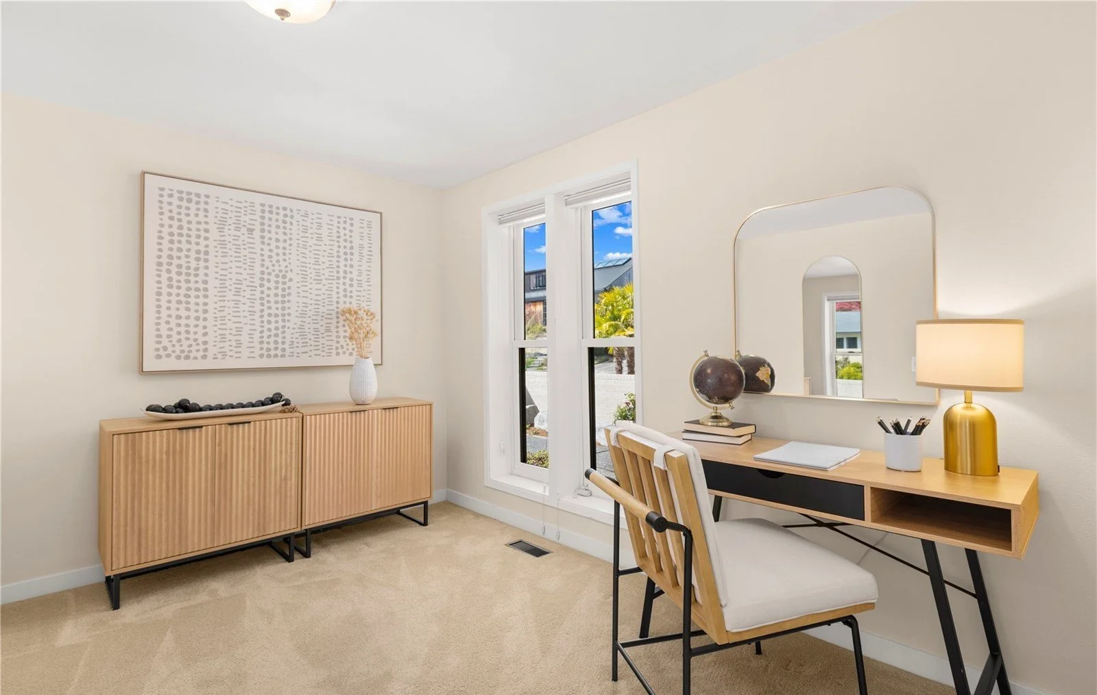 A bright, modern home office with a beige carpet, white walls, and a large window showing a sunny outdoor scene with trees and rooftops. It includes a wooden desk with a mirror, a white lamp, and decorative black globes, alongside a matching wooden c