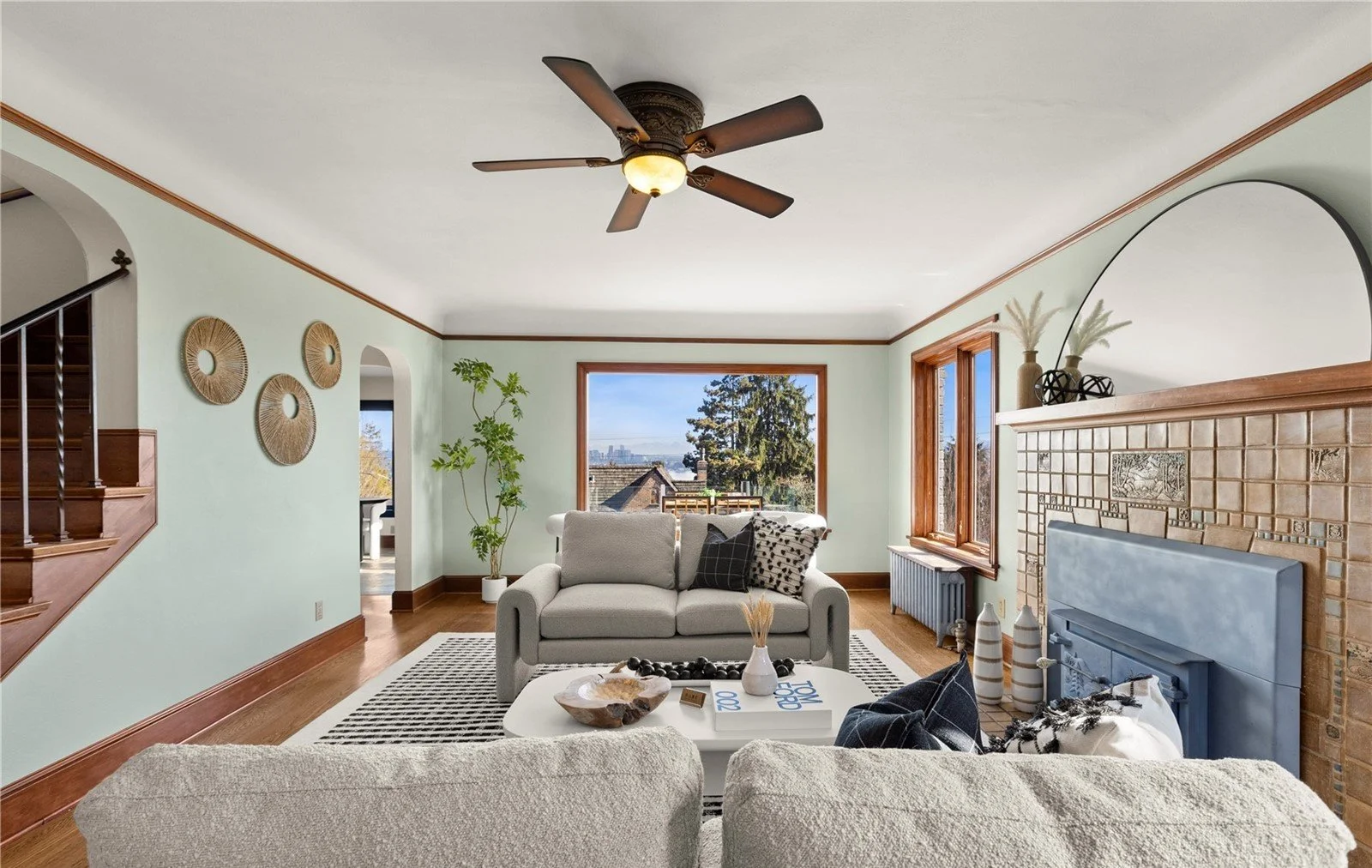 Living room with large window showing outdoor view, furniture including a gray couch, coffee table with decorative items, and fireplace with decor on mantel.