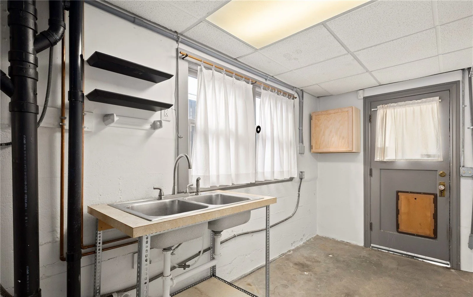 Laundry room with a double sink, white curtains, and pipes on the wall.