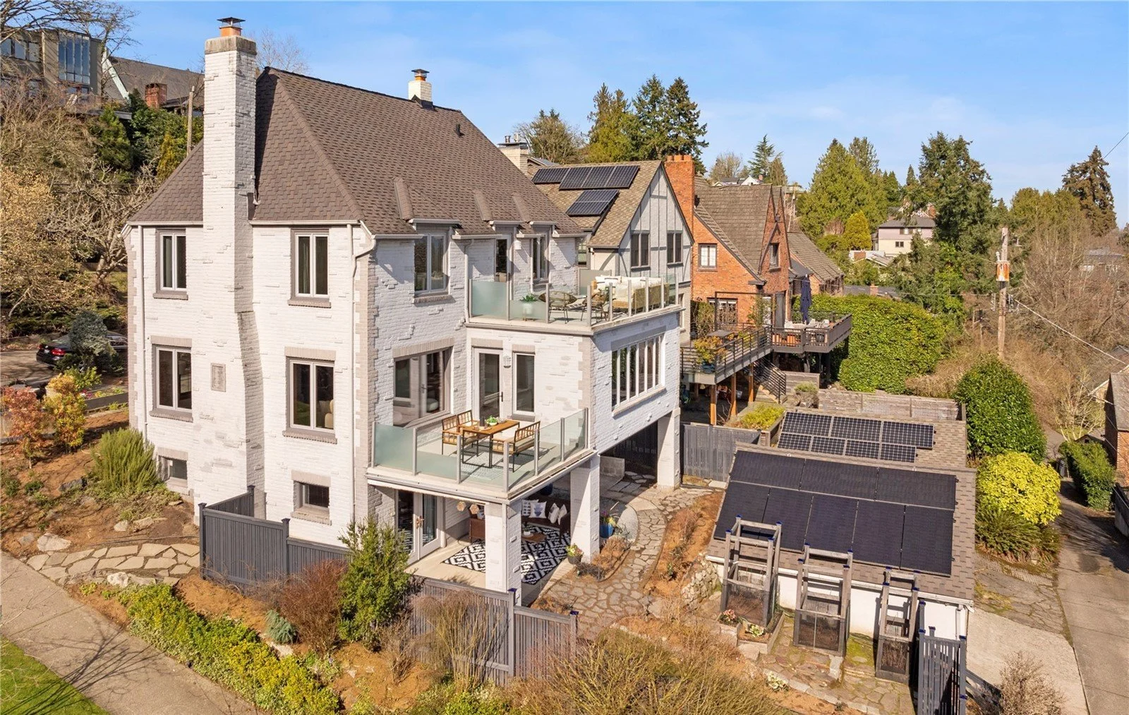 A multi-story house with modern balconies and a backyard with solar panels on the roof, surrounded by trees and neighboring houses, under a clear blue sky.