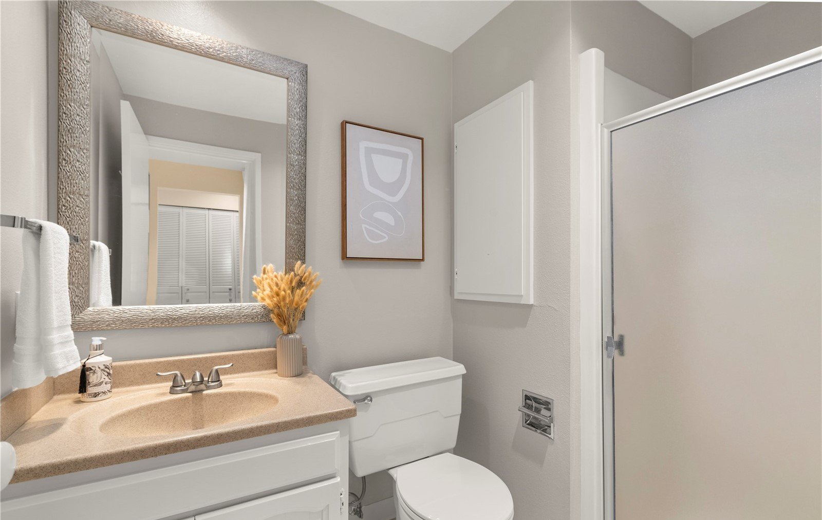 A small bathroom featuring a beige countertop with a built-in sink, a mirror with textured silver frame, a white toilet, a framed abstract art piece, and a shower with frosted glass door.