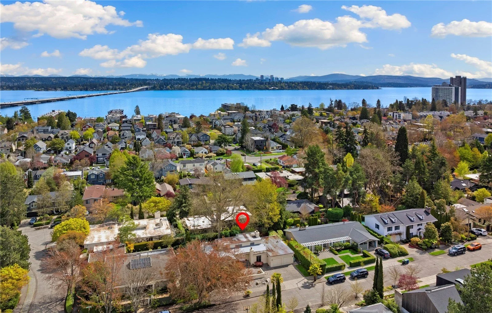 Aerial view of a suburban neighborhood near a large body of water with houses, trees, and a bridge, under a partly cloudy sky.