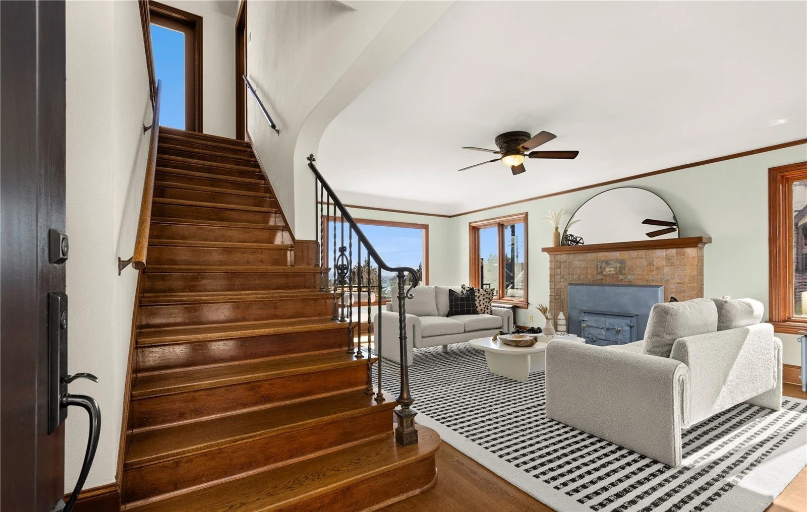 Living room with wooden staircase, white walls, large windows, a beige sofa, armchair, and a brick fireplace with a large round mirror above it.
