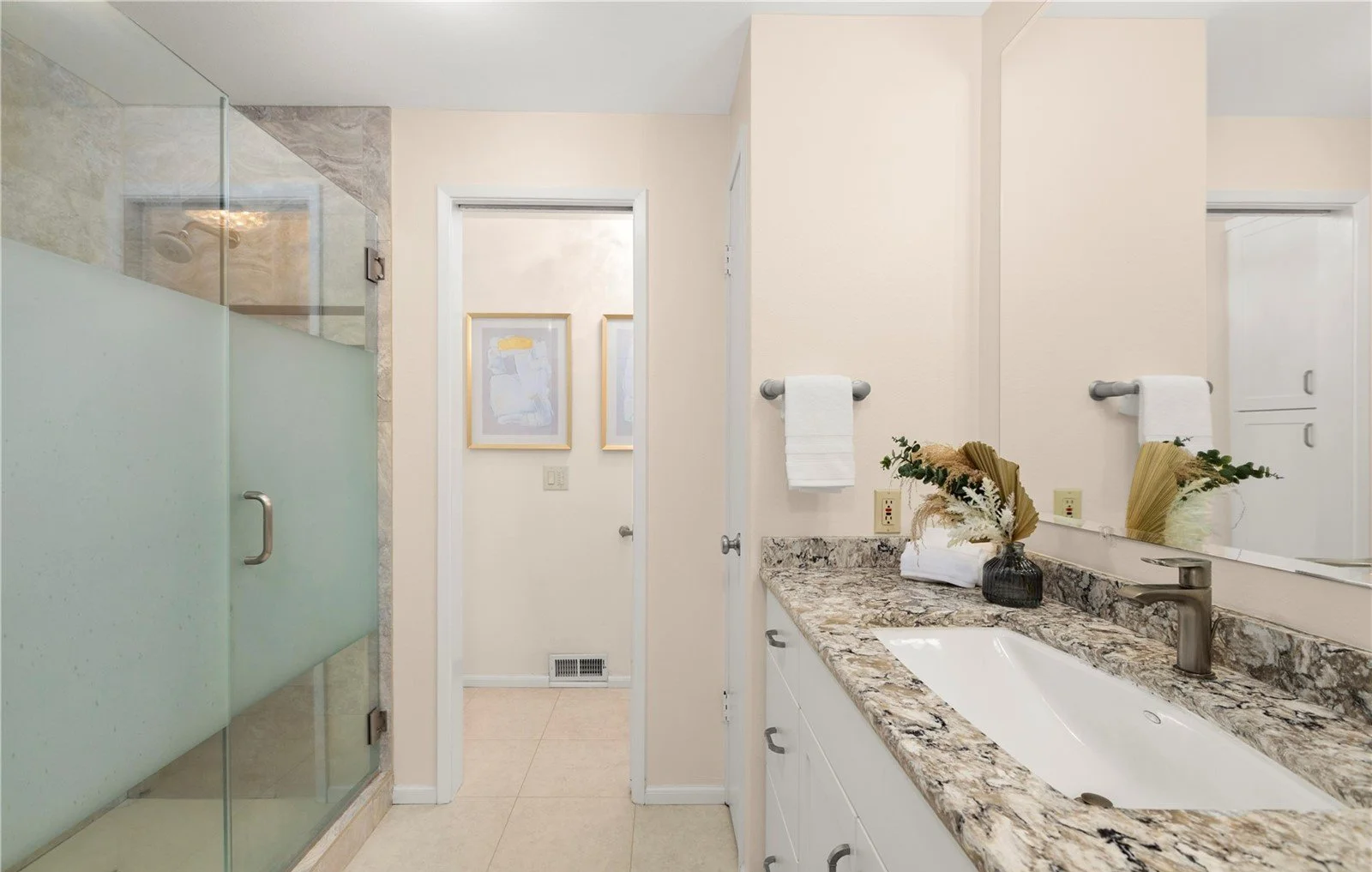 Bathroom with a glass shower enclosure, beige walls, a granite countertop with a white sink, a large mirror, and a small decorative vase with dried flowers.