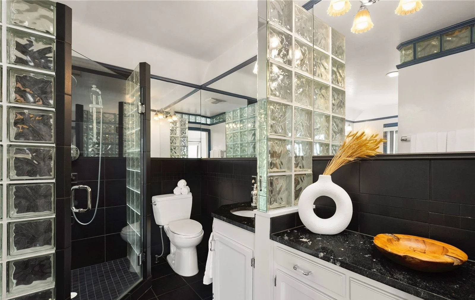Modern bathroom with glass block walls, black and white decor, and a shower stall to the left. The bathroom features a toilet with rolled towels on top, a white vanity with a black countertop, and decorative objects including a white vase with dried 
