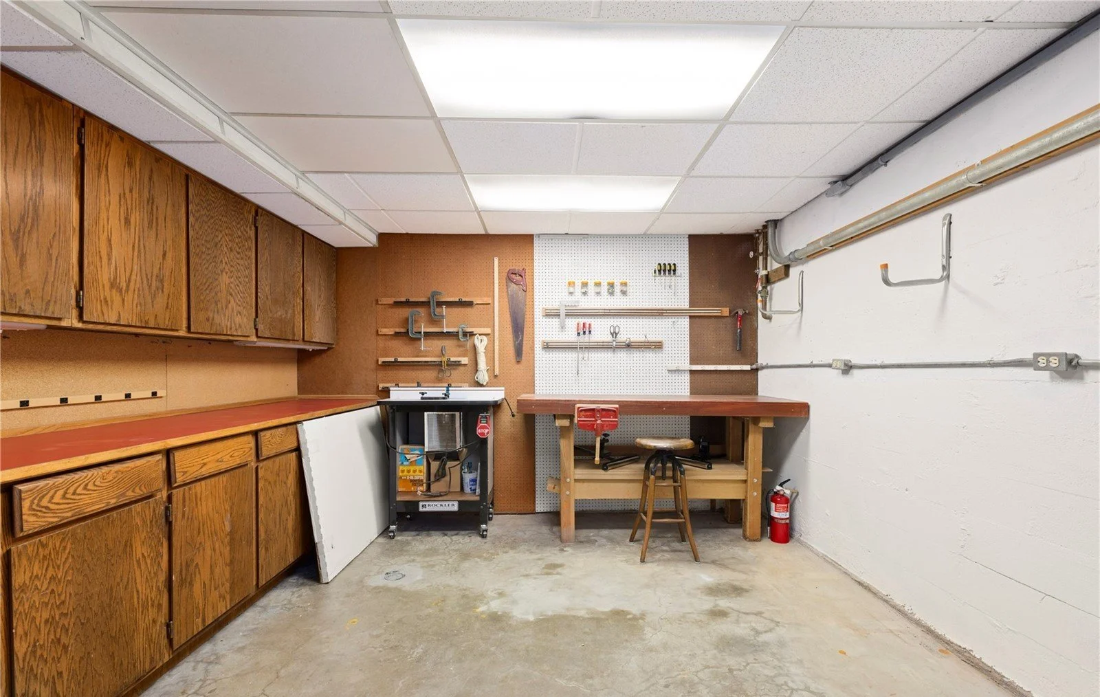 A garage workshop with wooden cabinets, a pegboard wall with tools, a wooden workbench with a stool, and a fire extinguisher.