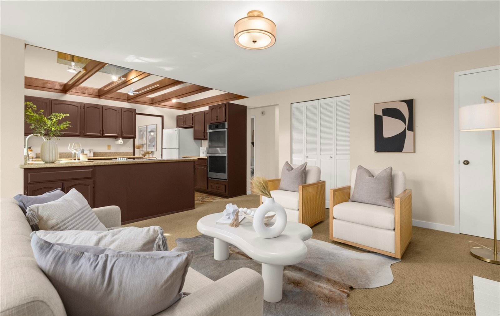 Living room with beige sofa and white coffee table, two armchairs with wooden armrests, wall art, and a floor lamp. Open kitchen with dark wood cabinets, countertops, and a ceiling with wooden beams.