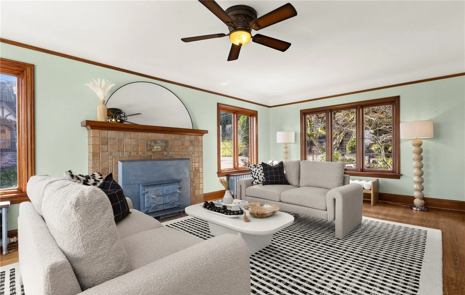 Living room with two beige sofas, black and white pillows, a white oval coffee table, a brick fireplace with a mirror above, wooden trim around windows and ceiling, hardwood floors, black ceiling fan, and large windows showing trees outside.