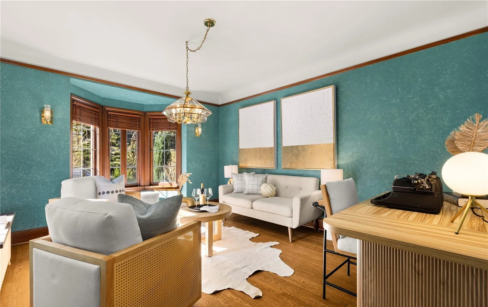 Living room with teal walls, wooden window frames, white sofa, armchair, coffee table, and two large abstract paintings in gold and white. Decor includes lamps, wine glasses, and a cowhide rug.