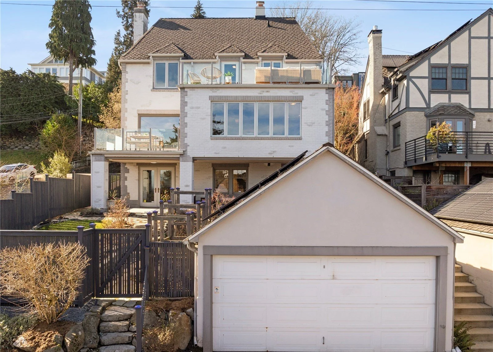 Multi-story house with white brick exterior, large upper-floor windows, balcony with outdoor furniture, and a detached garage in the front. Neighboring houses are visible on either side, with trees and sky in the background.