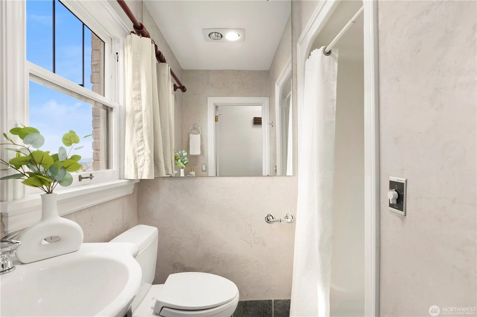 A small bathroom with a window, a mirror, a toilet with a plant on top, a towel rack, and a shower space with a curtain.