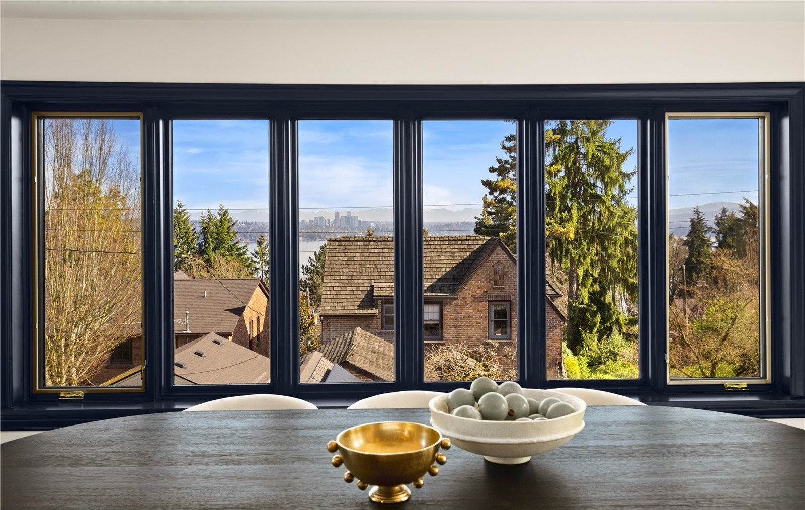 A view through a large window shows a suburban neighborhood with houses, trees, and distant city skyline under a blue sky with clouds.