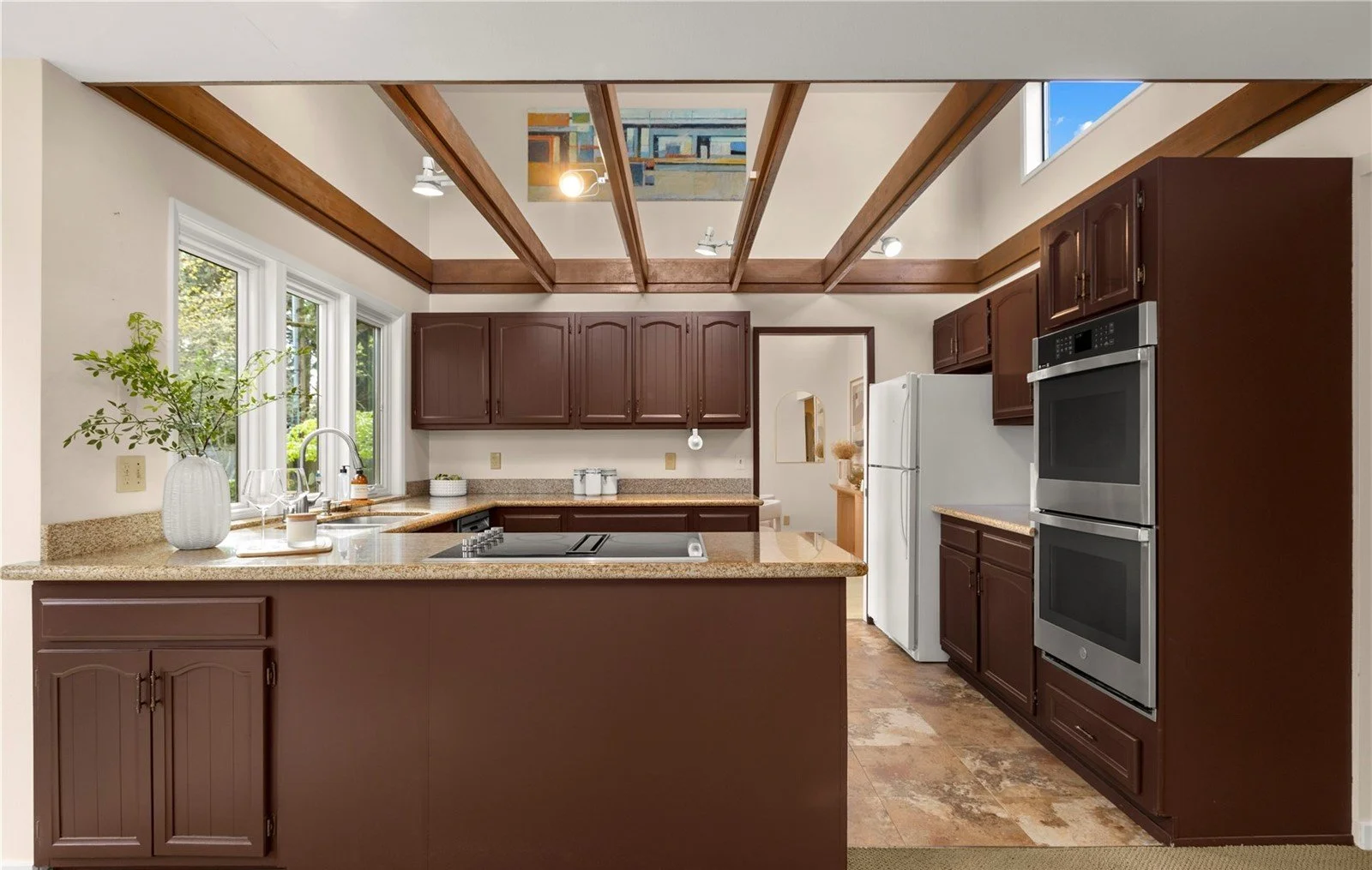 Kitchen with brown cabinets, beige granite countertops, white appliances, large window, and exposed wooden beams ceiling.