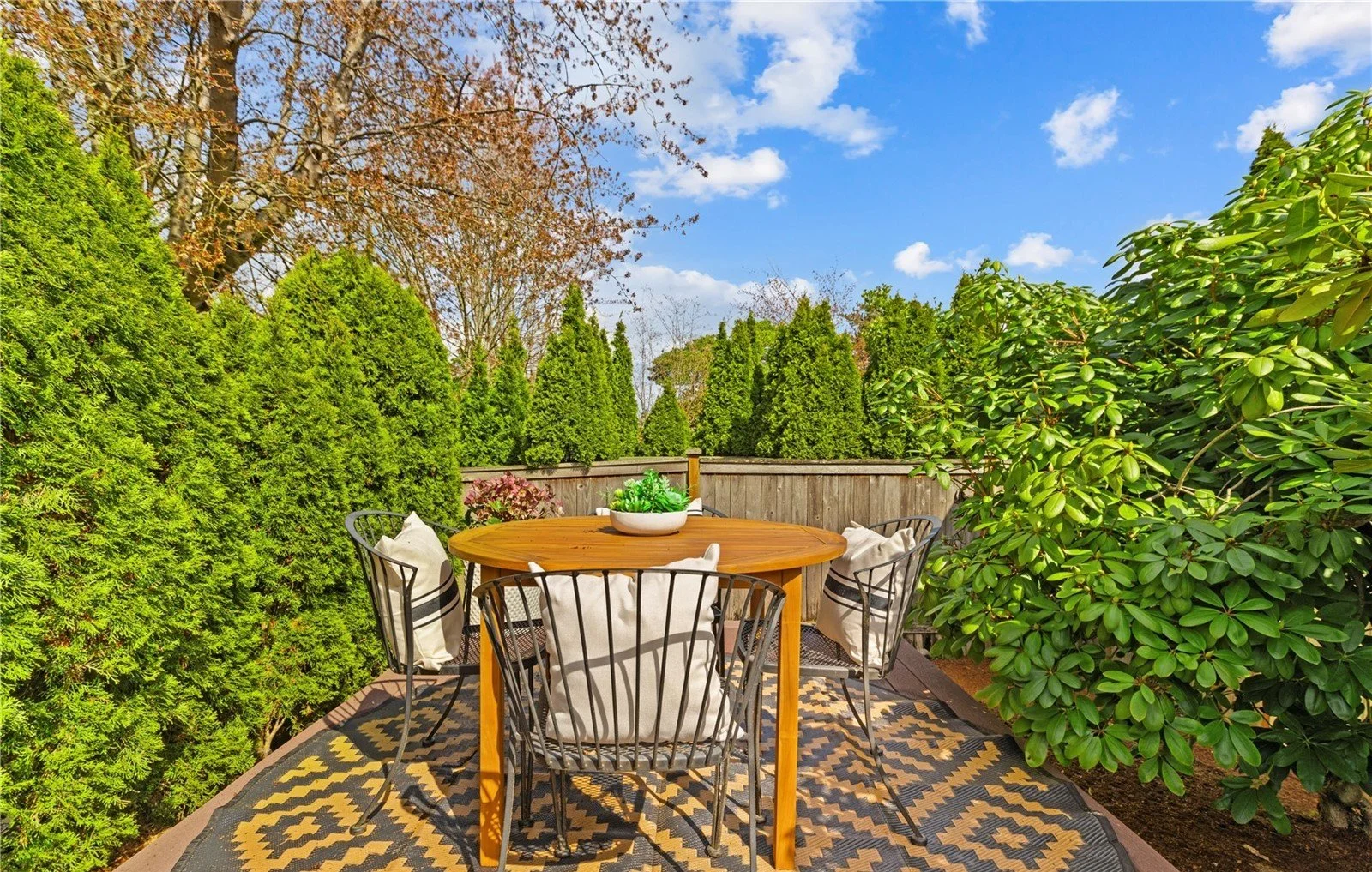 A backyard patio with a round wooden table surrounded by black metal chairs with white cushions. The table has a white bowl with green plants. The patio is enclosed by a wooden fence and lush green hedges and trees, under a partly cloudy blue sky.
