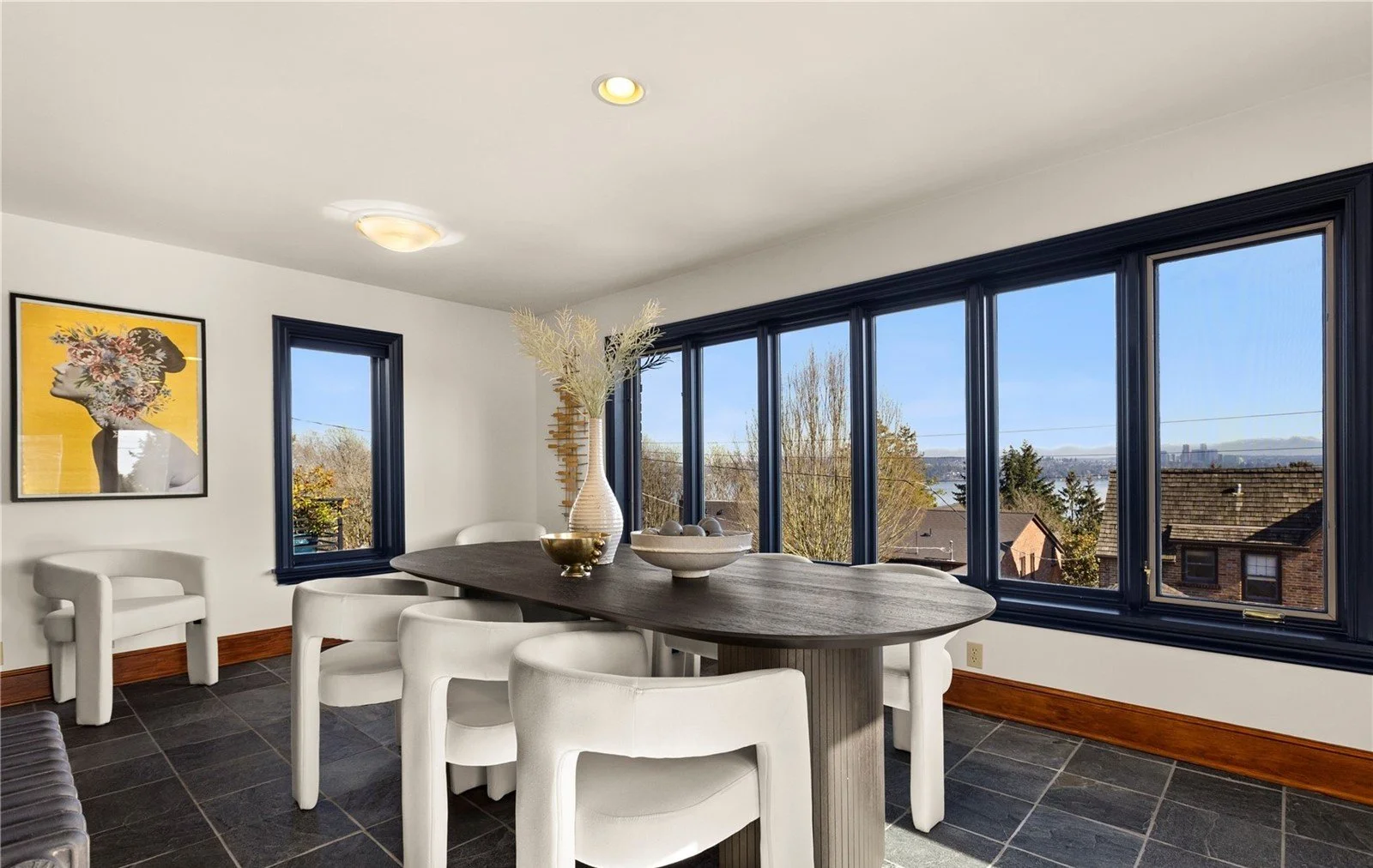 Dining room with a dark wood table, white chairs, large windows showing a view of trees and houses outside, and a white wall with a framed art piece.