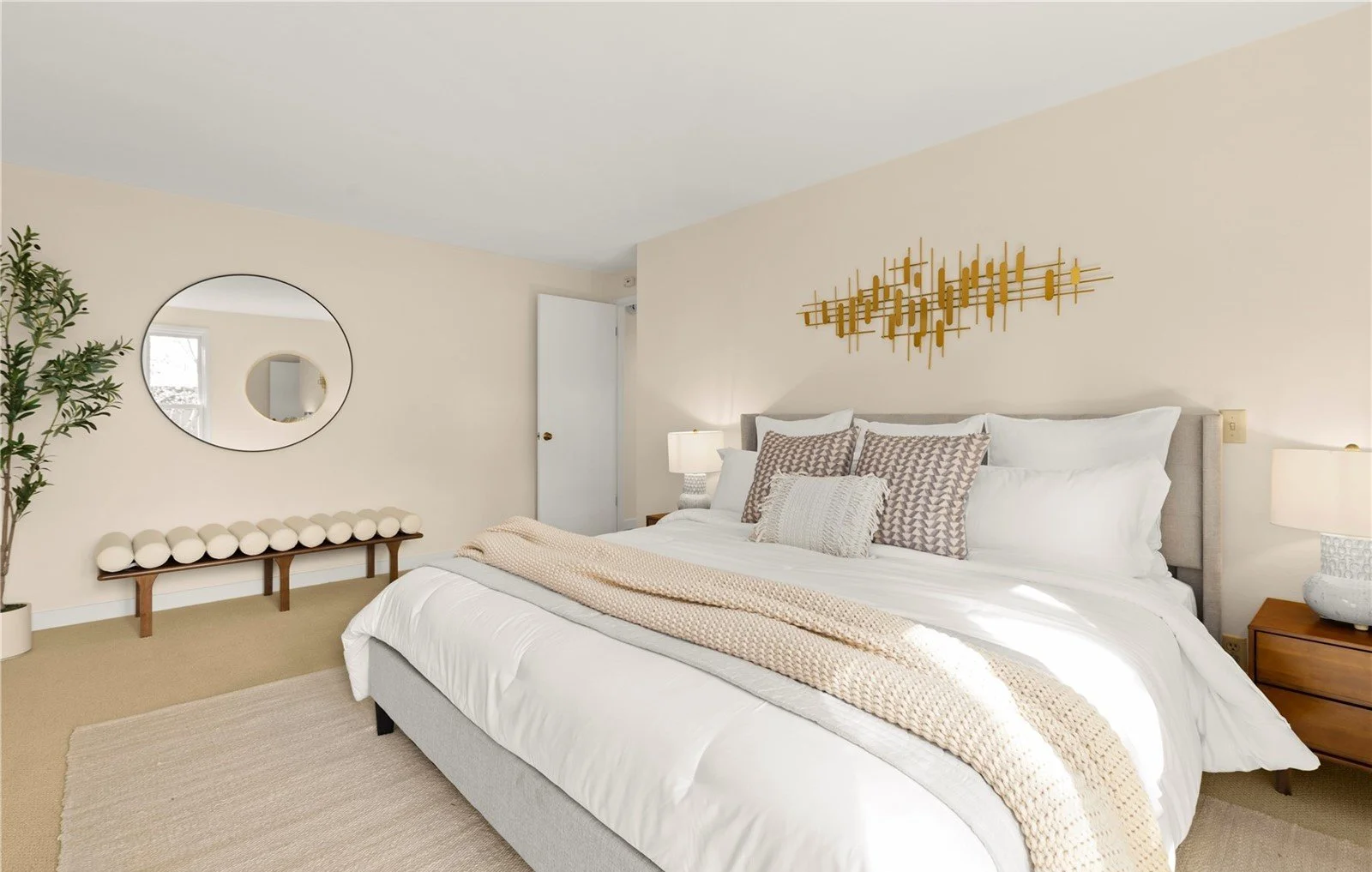 A bedroom with a large bed featuring white bedding, beige and patterned pillows, and a beige knit throw blanket. There are two bedside tables with lamps, a round mirror on the light-colored wall, a bench with decorative balls, a tall potted plant, an