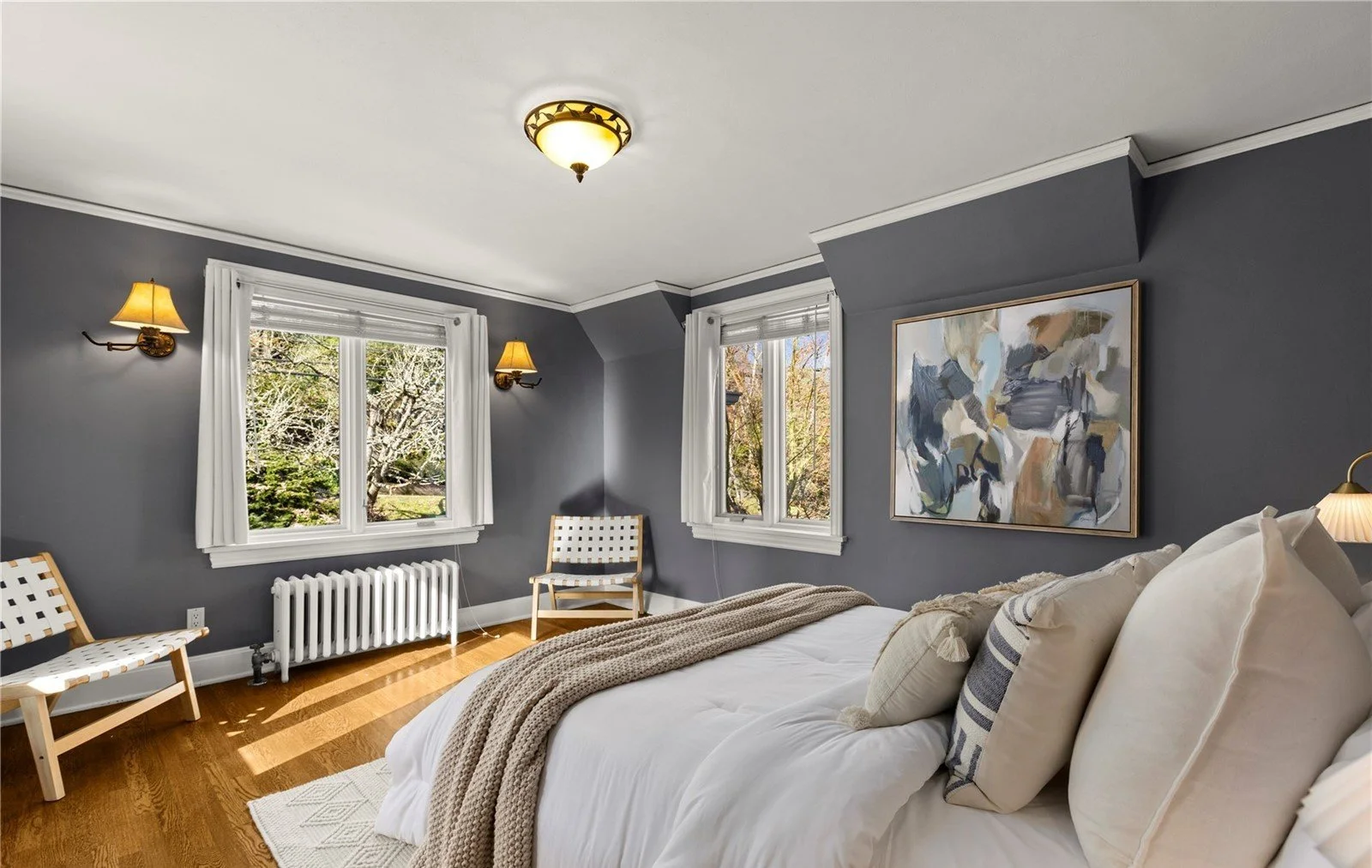 A bedroom with gray walls, two windows with white curtains, two wall lamps, hardwood floors, a bed with white bedding and beige pillows, a tan throw blanket, an abstract painting on the wall, two white chairs with wooden frames, and a ceiling light f