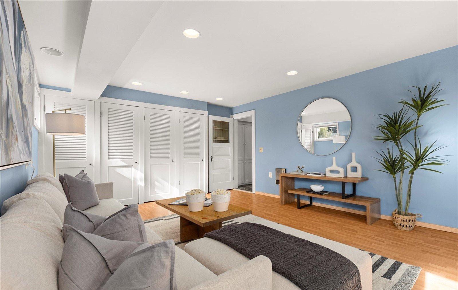 Living room with a beige sofa, wooden coffee table, and blue accent wall. There are white louvered closet doors, a round mirror, a wooden bench with decor, and a potted plant. Recessed lighting and natural light are present.