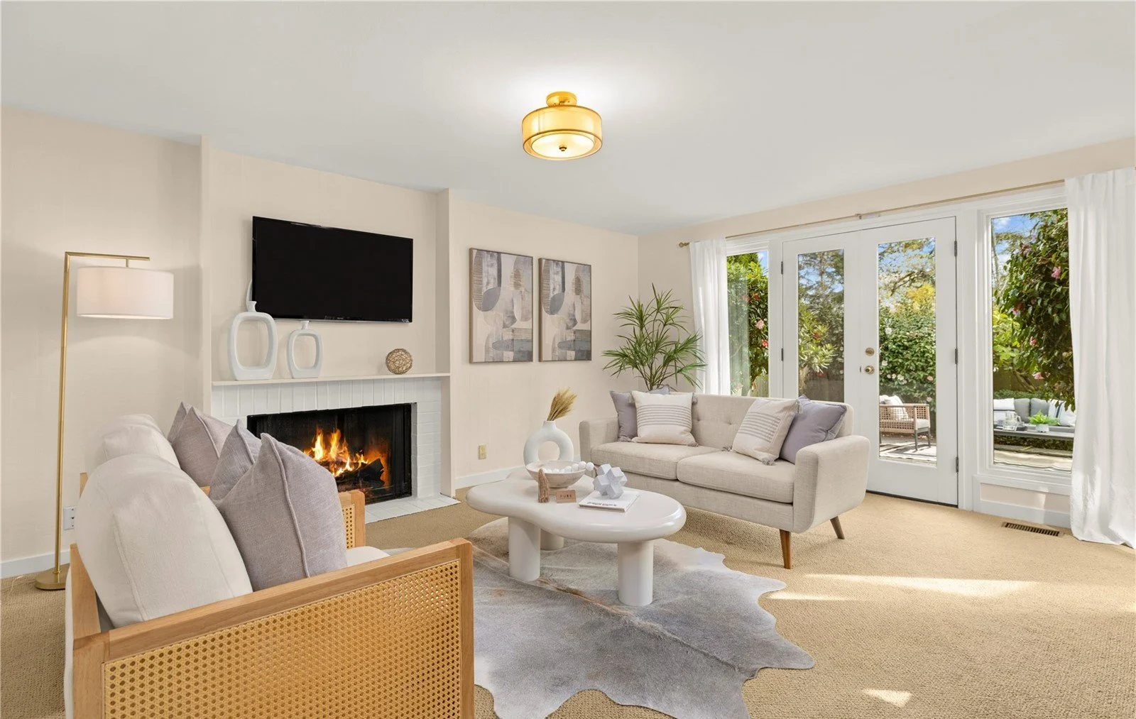 Bright living room with beige walls, a fireplace, a wall-mounted flat-screen TV, a white sofa, a rattan armchair, a white coffee table, and large glass doors opening to a garden with outdoor seating.