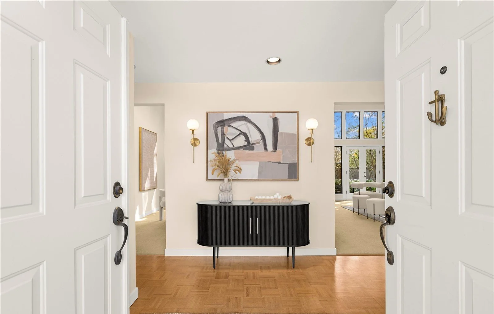 View through open white double doors into a modern living space with a black sideboard, abstract art, and wall-mounted lamps.