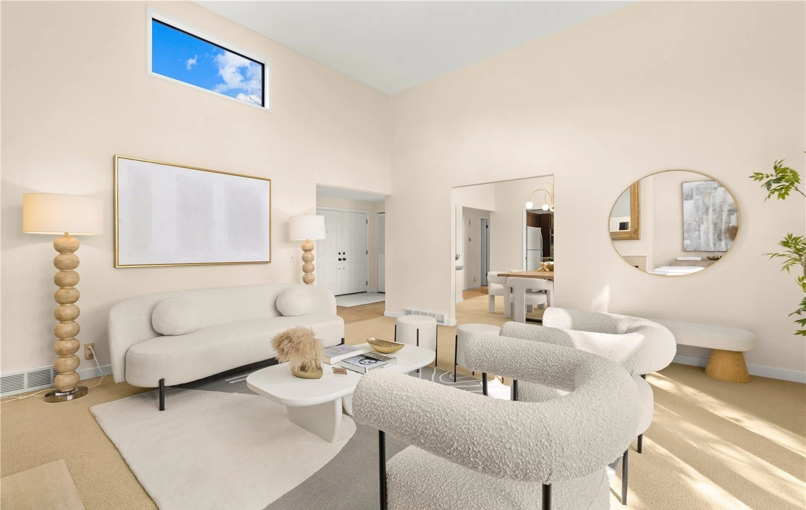 Modern living room with white furniture, decorative lamps, a large abstract painting, and a round mirror on the wall. There is a view into the dining area and kitchen, with a high window showing blue sky and clouds.