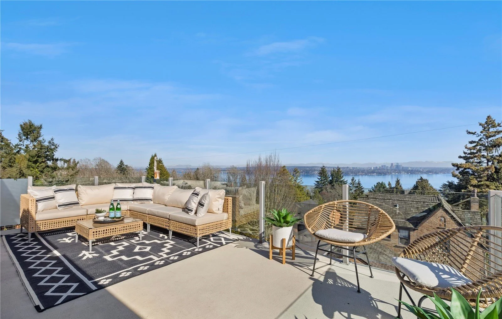Outdoor balcony with wicker sectional sofa, patterned black and white rug, two rattan chairs with white cushions, small potted plant, and bottles of drinks, overlooking a lake and city skyline under a clear blue sky.