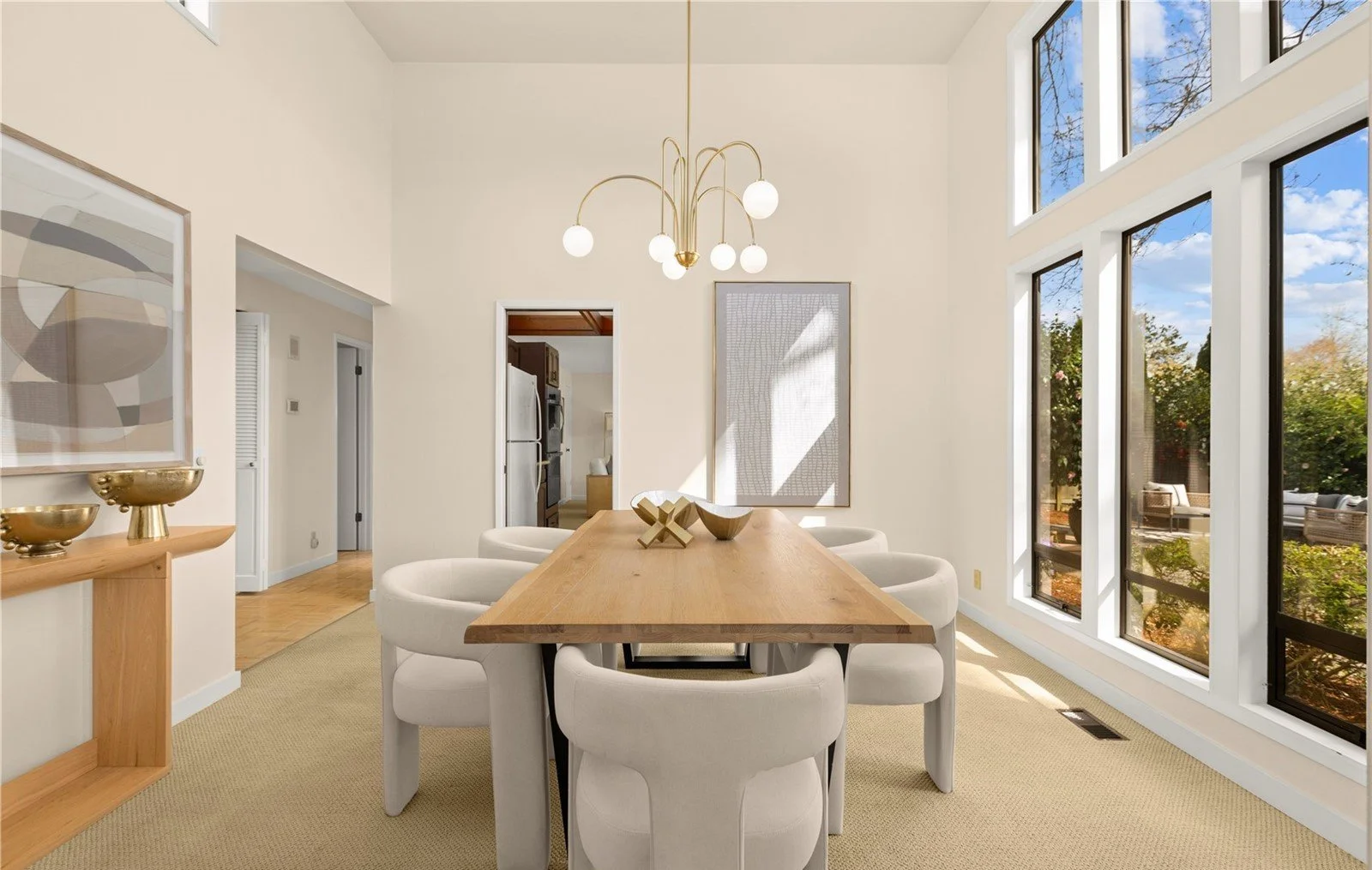 Bright dining room with round white chairs, a wooden table, large windows with a view of trees, and modern art and decor.