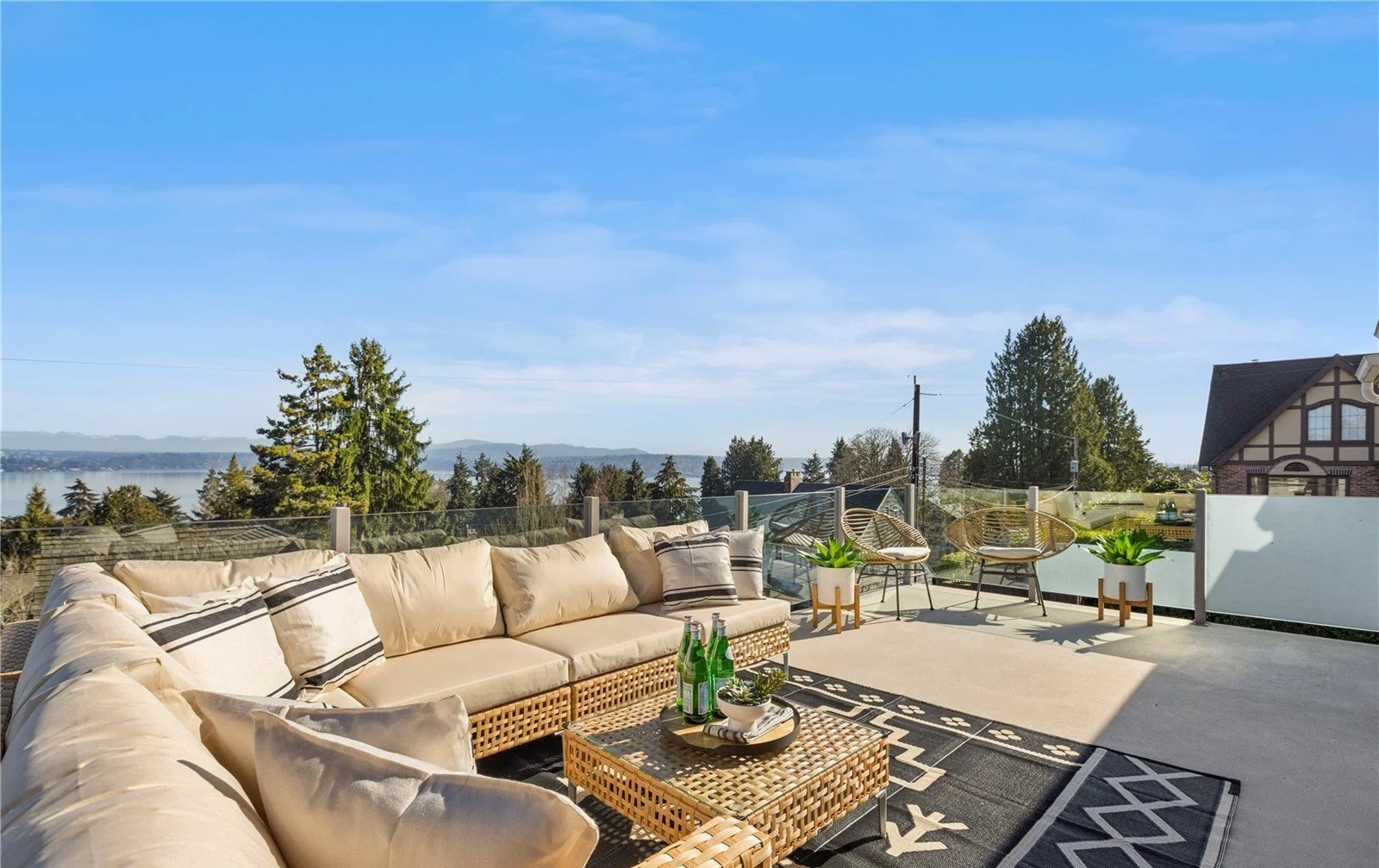 Outdoor rooftop patio with beige sectional sofa, black and white pillows, wicker coffee table with drinks, two wicker chairs, potted plants, and scenic view of trees and water under a clear blue sky.