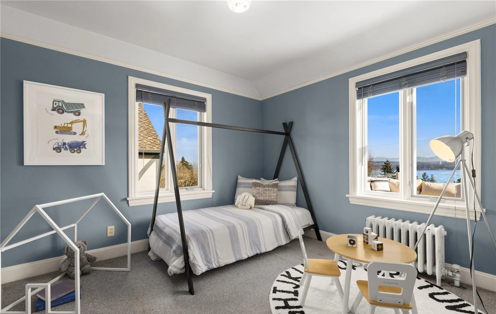 A cozy children's bedroom with blue walls, a bed with striped bedding, a small table with chairs, a house-shaped shelf, and large windows showing a scenic view outside.