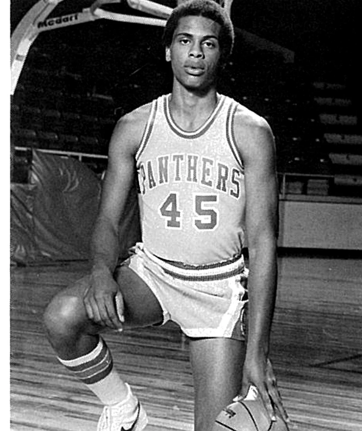 A black and white photo of a young man in a basketball uniform, holding a basketball and kneeling on a basketball court. He is wearing a jersey with the number 45 and the word 'PANTHERS' on it.