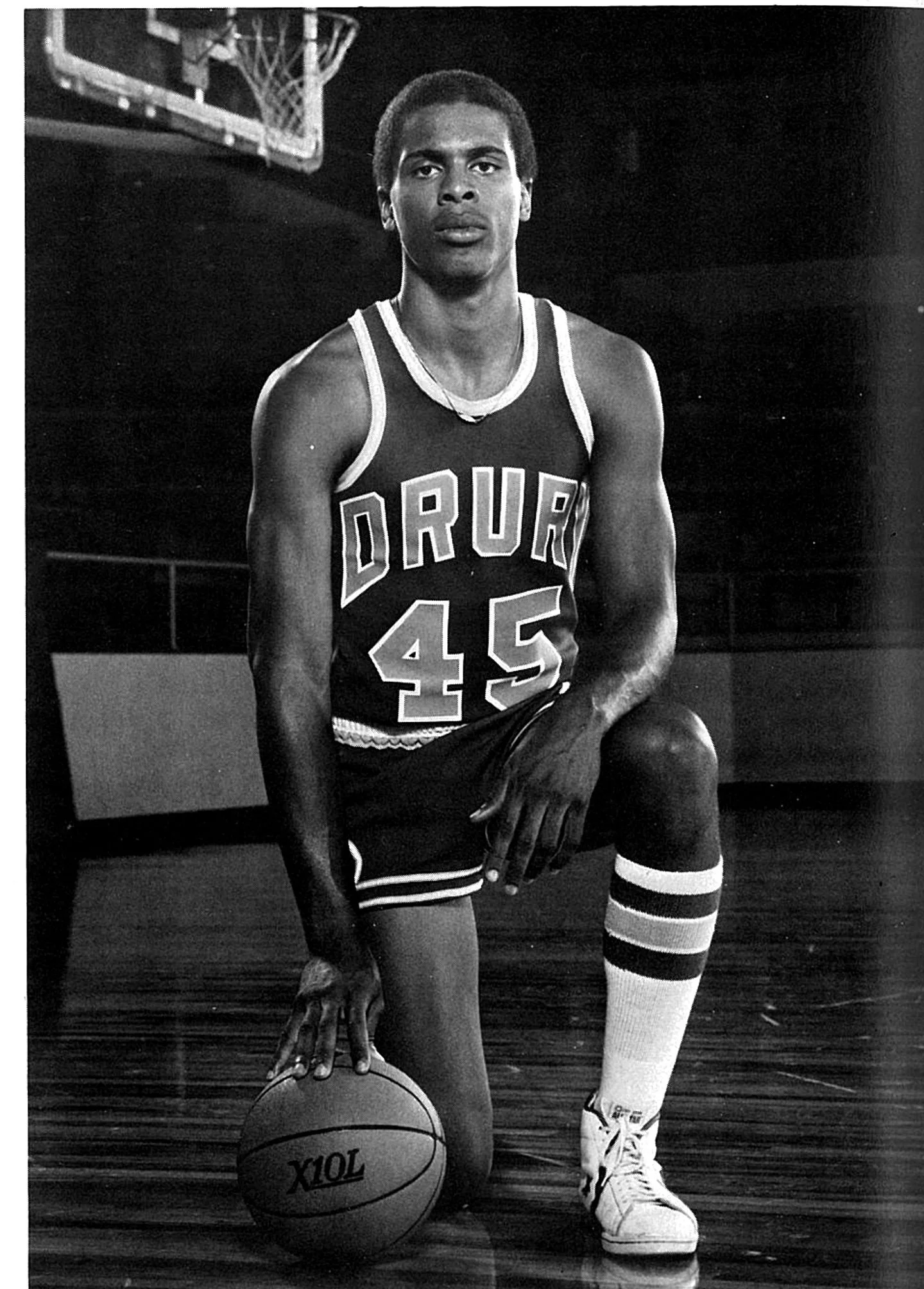 Black and white photograph of a young African American male basketball player in a vintage jersey with the name "DURKEE" and the number 45. He is kneeling with one knee on a basketball, and the other foot on the ground. He wears striped knee-high socks and sneakers, with a basketball hoop visible in the background.