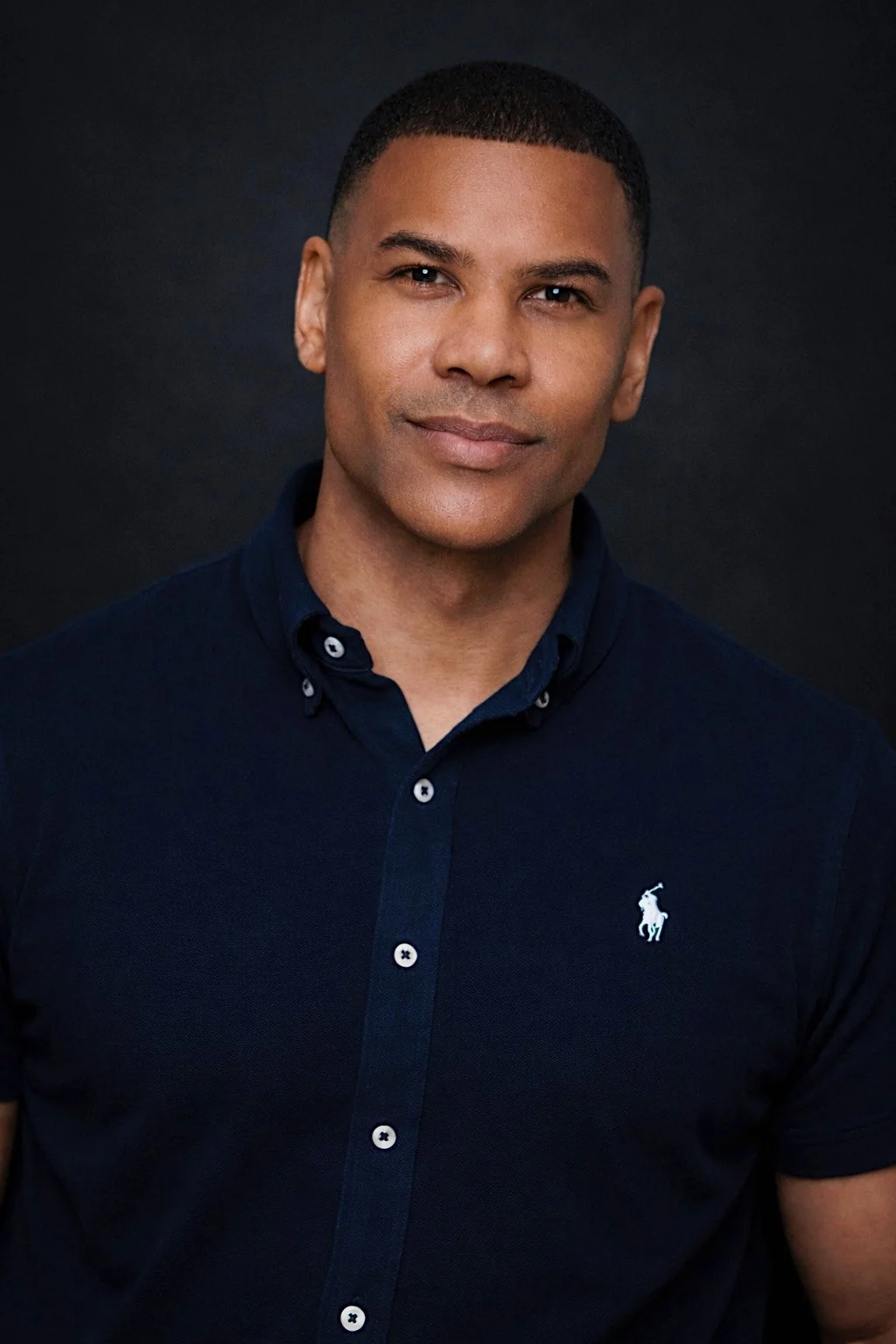 A man with short hair and brown skin wearing a navy blue polo shirt with a small white logo on the chest, posing against a black background.